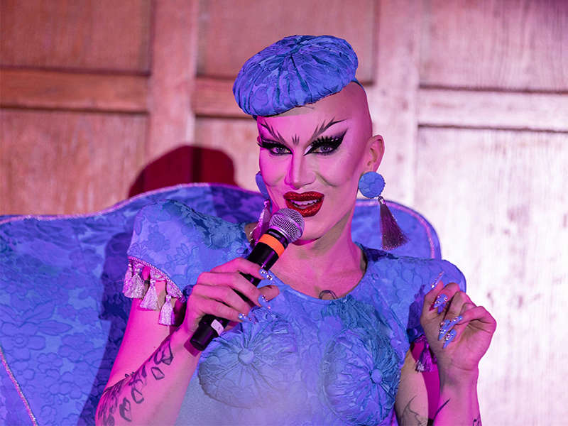 A person with red lipstick, a shaved head, and a bright indigo dress and cap speaks into a microphone.