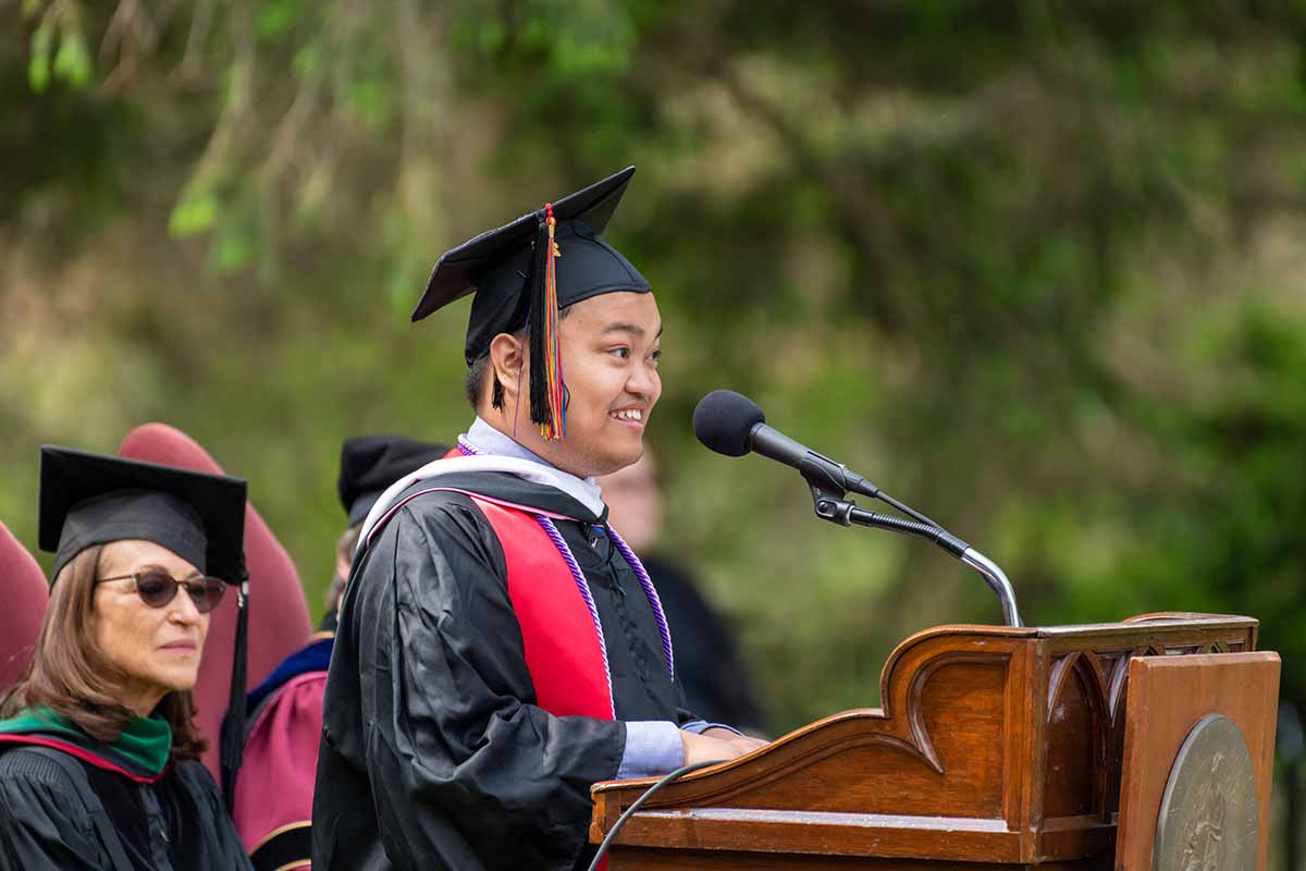 A person with short black hair, a graduation, cap, and a robe, stands in front of a podium, facing to the left, speaking into a microphone.