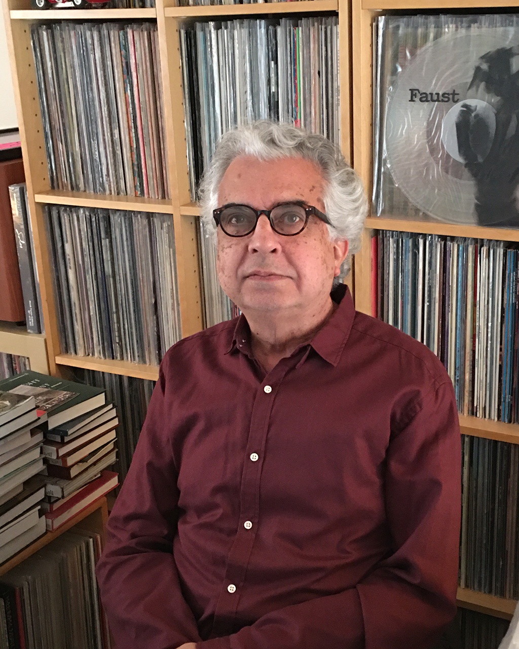 A man with dark glasses and gray hair sitting in front of a bookcase filled with vinyl records.
