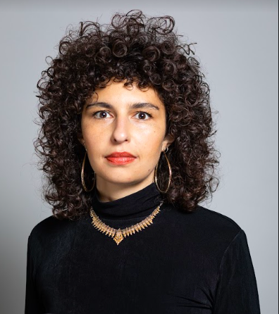 Person with dark curly hair and a black shirt with a necklace looking at the camera.