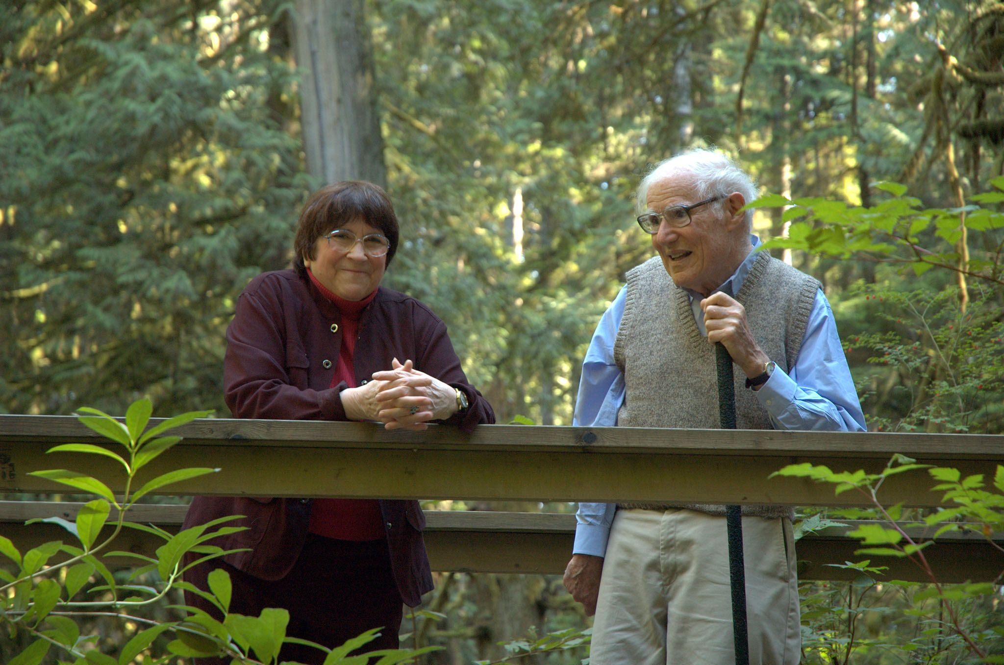 An elderly man and woman standing together on a wooden walkway in a lush, forested area, smiling and talking.