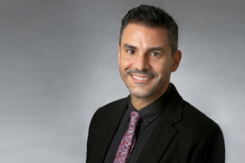 Carlos Alamo wearing a black shirt and red patterned tie against a gray background.