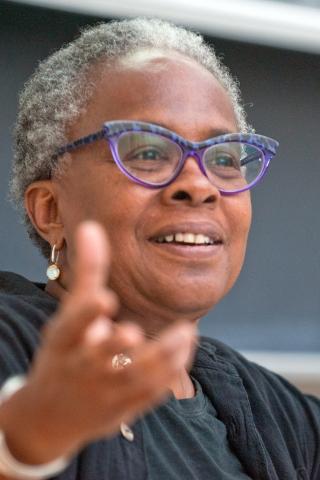 Diane Harriford wearing a black shirt and purple eyeglasses with arm extended against a dark background.