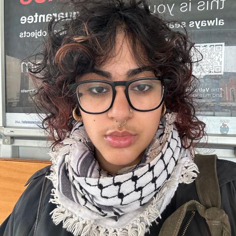 Person with black curly hair, dark glasses, scarf, and black jacket with both a nose ring and stud..