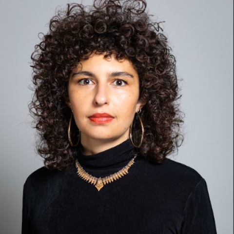 Person with dark curly hair and a black shirt with a necklace looking at the camera.