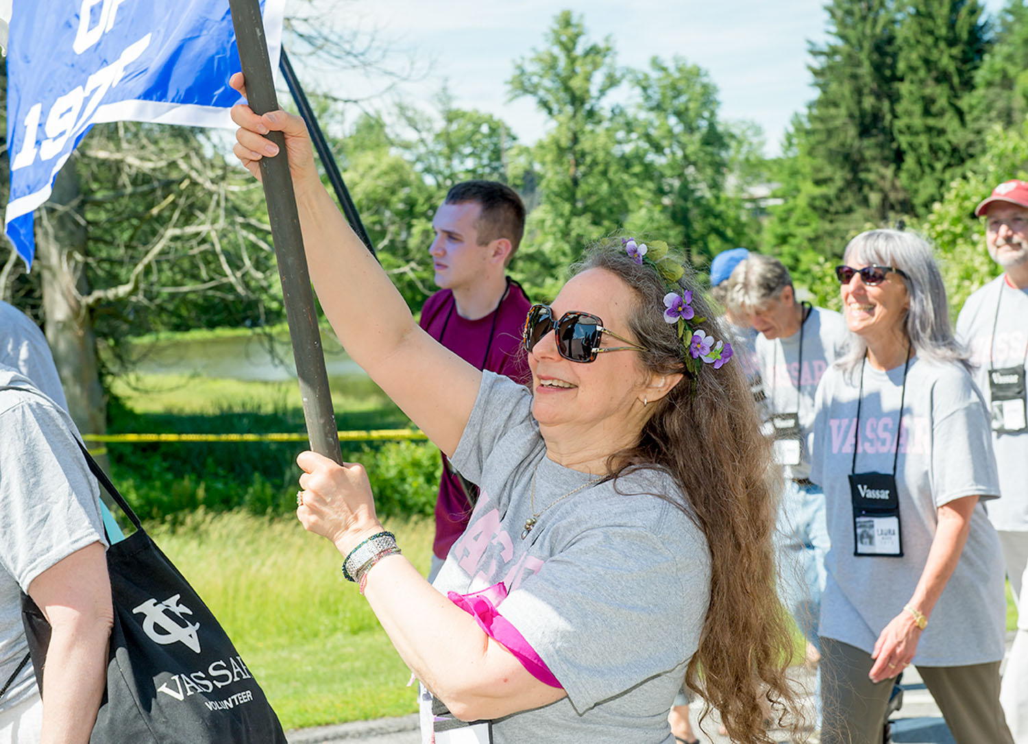 People walking in a group, with a person with long hair and flowers in it holding a banner that says Class of 1975 on it.