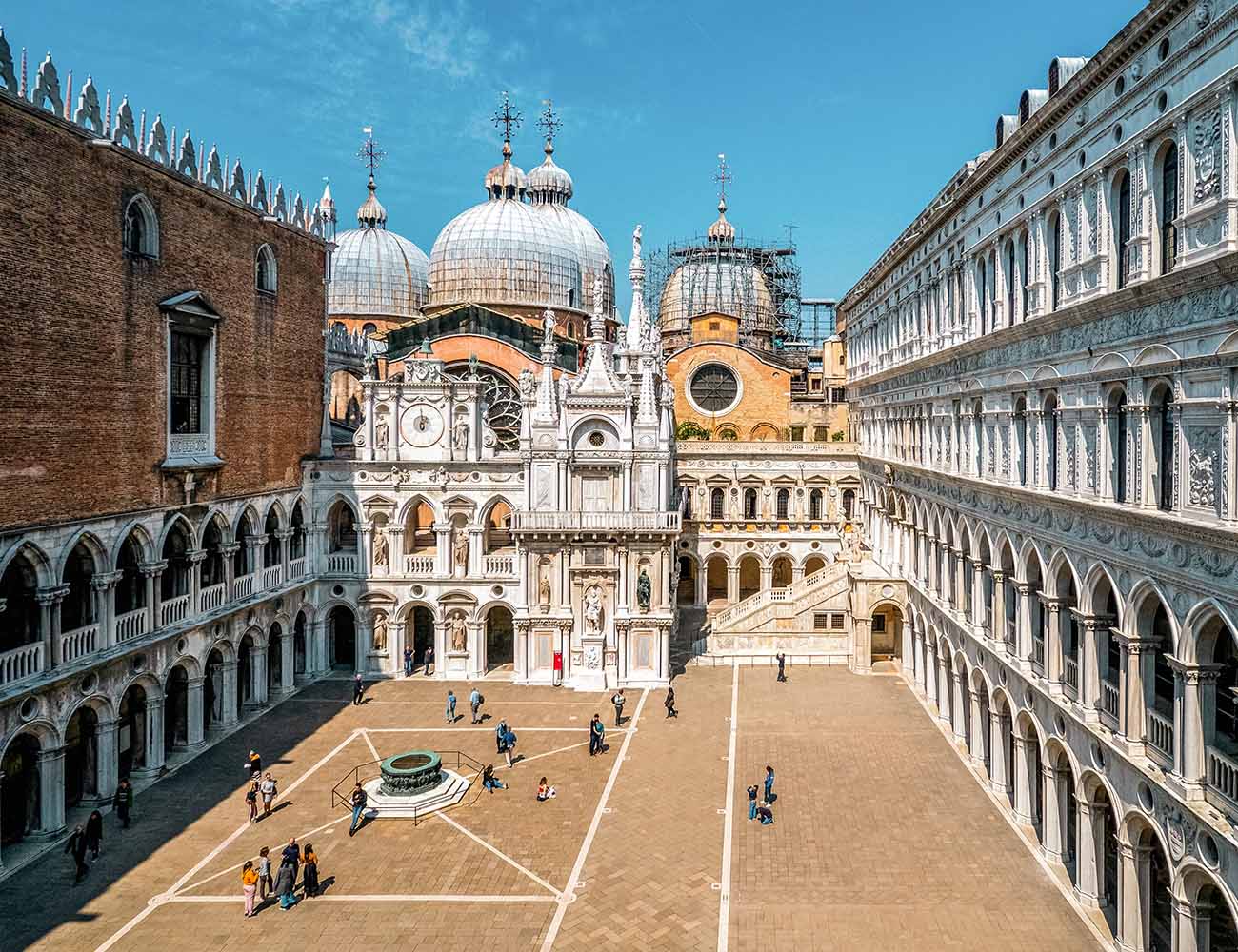 A large courtyard with several stories of brick or colonnaded walls on three sides, with the round domes of the church of San Marco in the background.