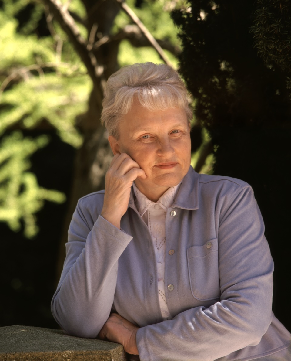 A person with short gray hair wearing a light gray jacket over a white shirt leans on stone block in front of green trees.