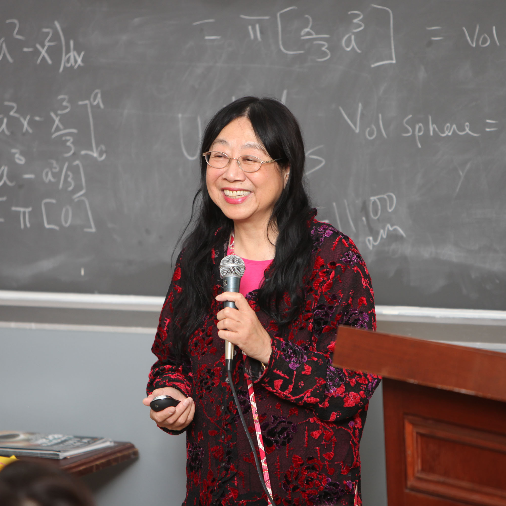 A person with long black hair wearing a red and black patterned jacket stands in front of a blackboard with equations written on it, holding a microphone and smiling.