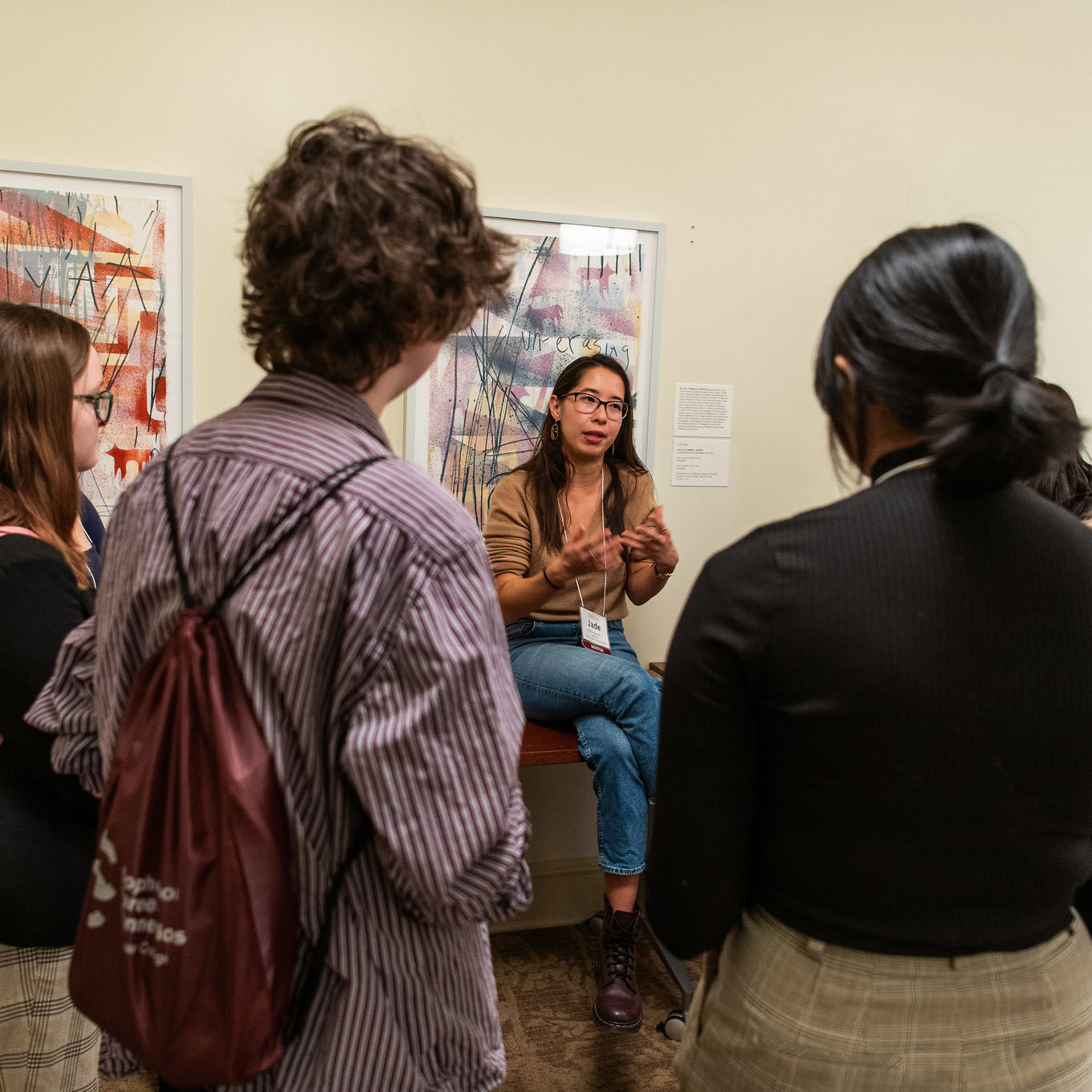 A person with long black hair sitting on a table in front of a wall talks to several students who stand in a semi-circle with their backs to the viewer.