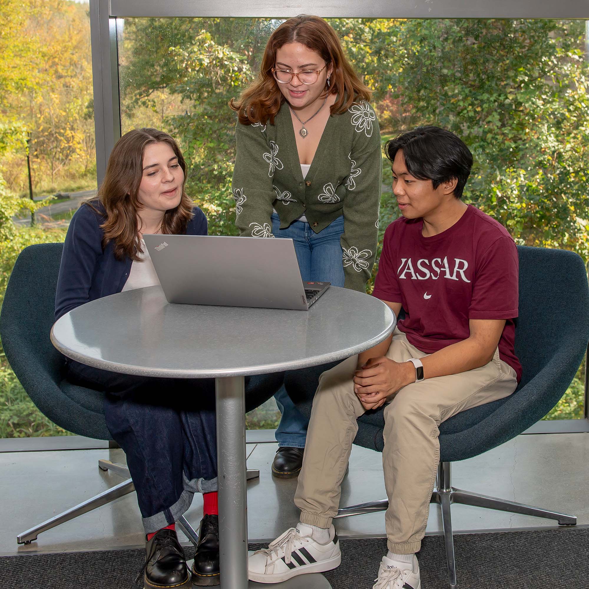 Two people sit in chairs at a small round table with a laptop computer on it,  in front of a large glass window, while another person stands between them in front of the computer, pointing at something on the screen.