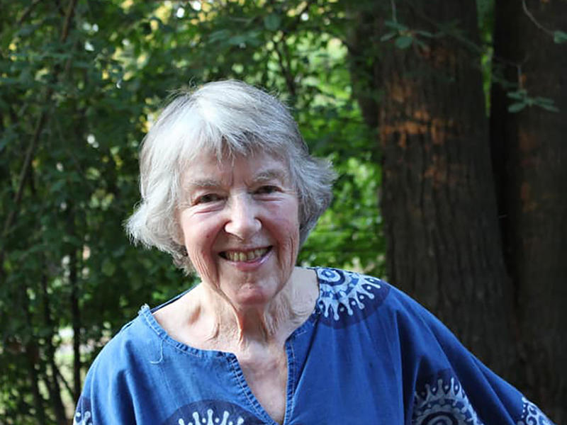 A person with mid-length gray hair and a blue patterned shirt smiles at the viewer. The person is outside, with trees and a tree trunk visible in the background.