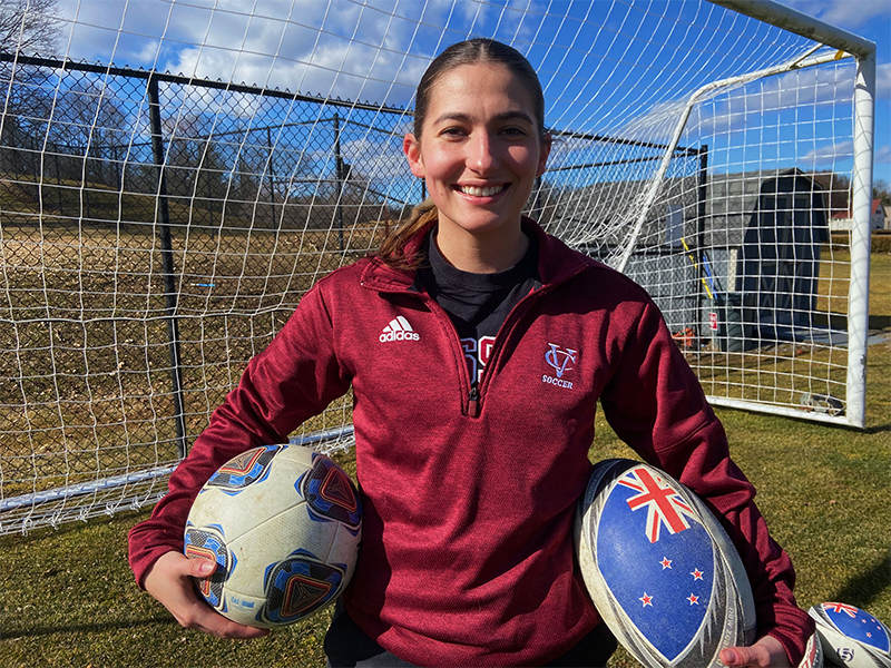 A person with long brown hair and a maroon athletic sweater stands outside in front of an athletic net, holding a ball in one hand and a helmet in the other.
