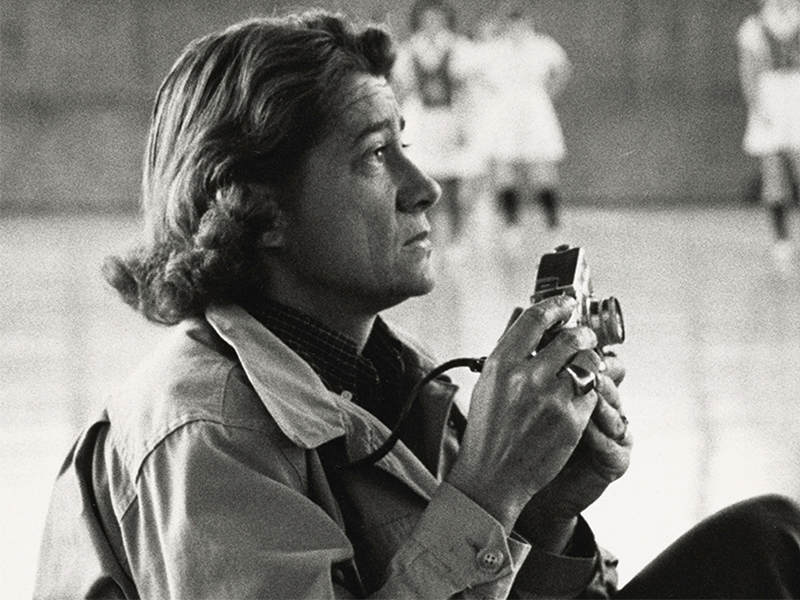 A black-and-white historical photo of a person with long hair, holding a camera and looking up.