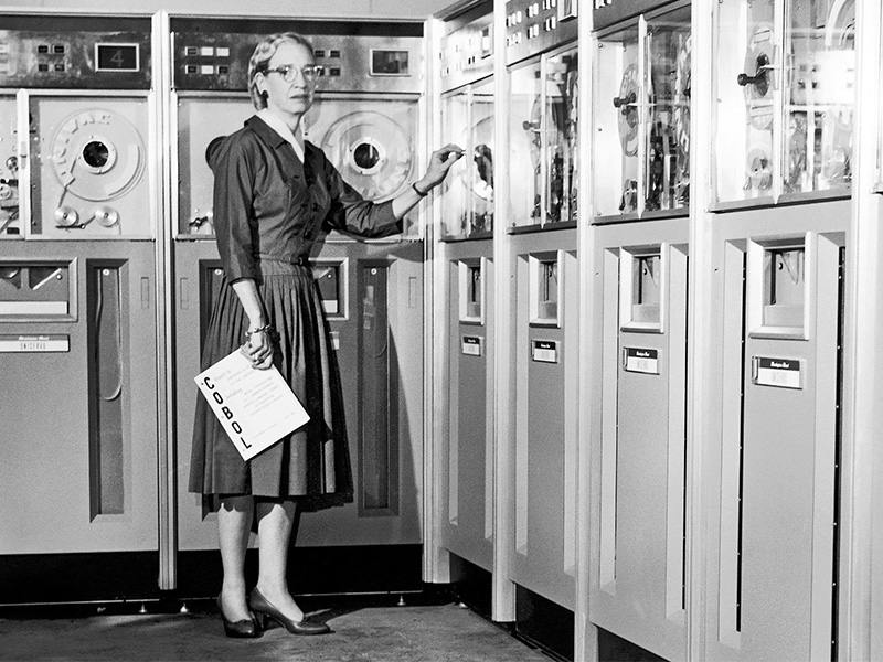 A monochrome historical photo of Grace Hopper, a person wearing a long dress and glasses, standing in front of old computer cabinets.