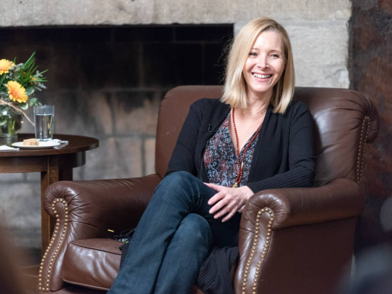 A photo of Lisa Kudrow ’85, a person with long blond hair, a dark shirt, and jeans. Kudrow is sitting in a brown leather chair near a fireplace.