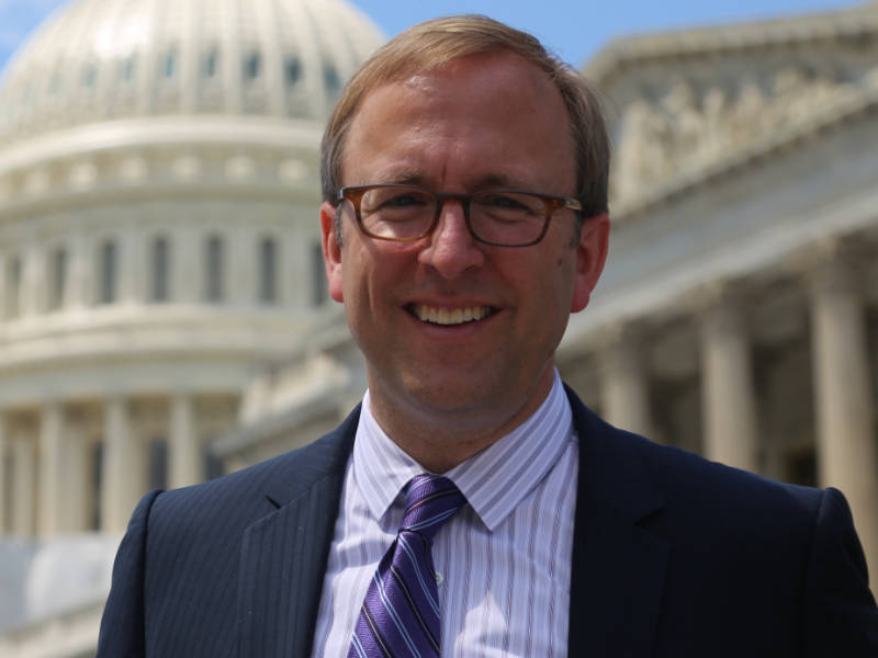 A photo of Jonathan Karl ’90, a person with short light-colored hair, glasses, and a dark suit. Karl is standing in front of the Capitol building, a classic building with a dome.