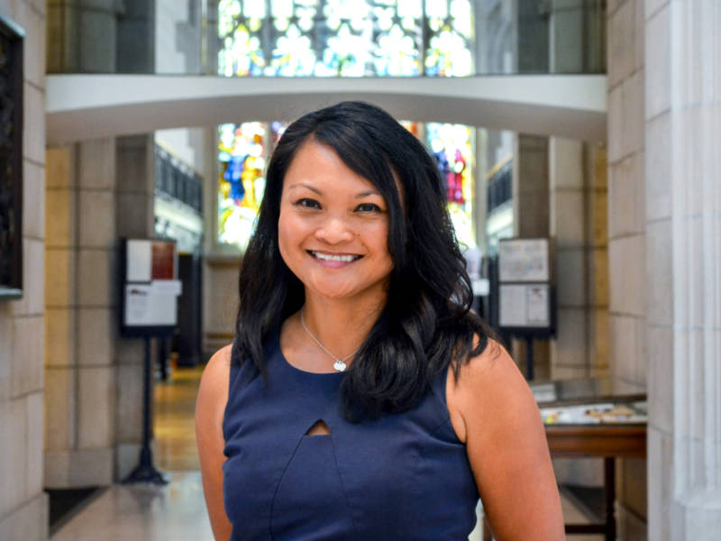 A photo of Michelle Tugade ’95, a person with long dark hair and a blue dress. Tugade is standing in the Vassar Library, a large stone building with a stained glass window in the background.