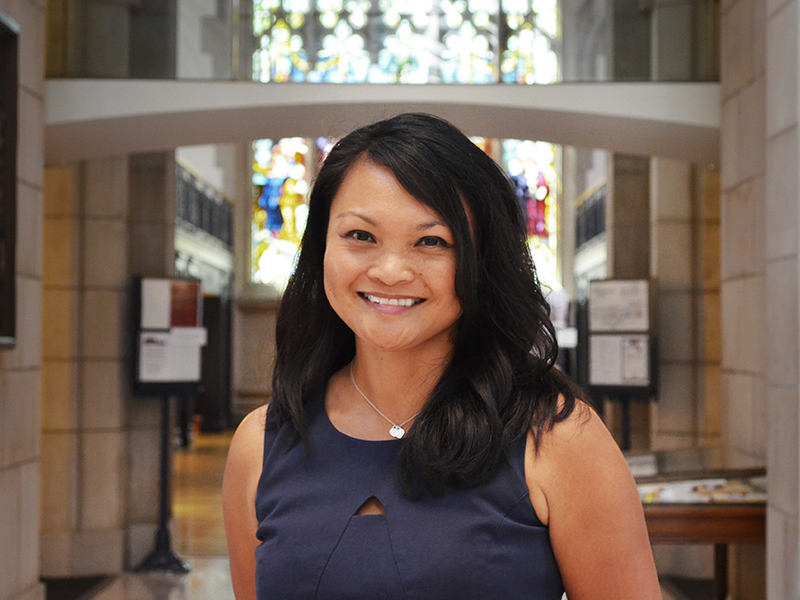 A photo of Michele Tugade ’95, a person with long black hair and a blue dress. Tugade is standing in the Vassar Library, a large stone building with a stained glass window behind her.