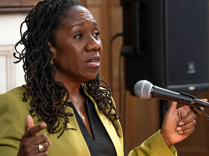 A photo of Sherrilyn Ifill ’84, a person with long black hair and a green dress. Ifill is speaking in front of a microphone.