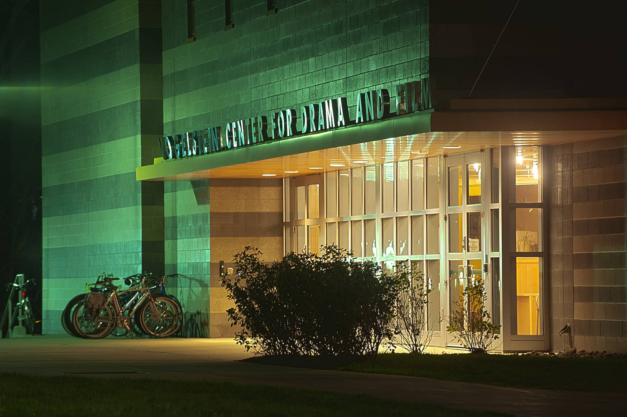 A nighttime view of the glass-walled entrance to a large stone and brick building, with the name of the building above the entrance, the Vogelstein Center for Drama and Film.