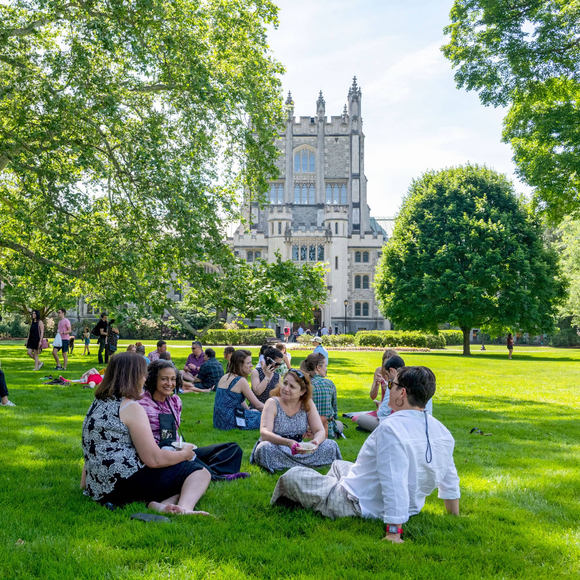 People seated on the lawn in front of Thompson Library, a large, stone building with stained-glass windows and classical architecture, on the Vassar campus.