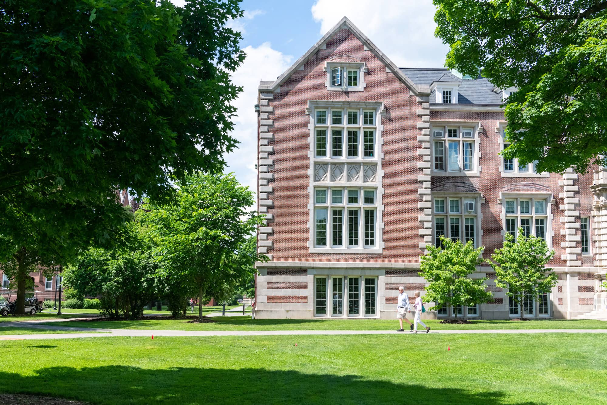 Two people walking in front of Rockefeller Hall, a large red brick building with many windows on the Vassar campus.