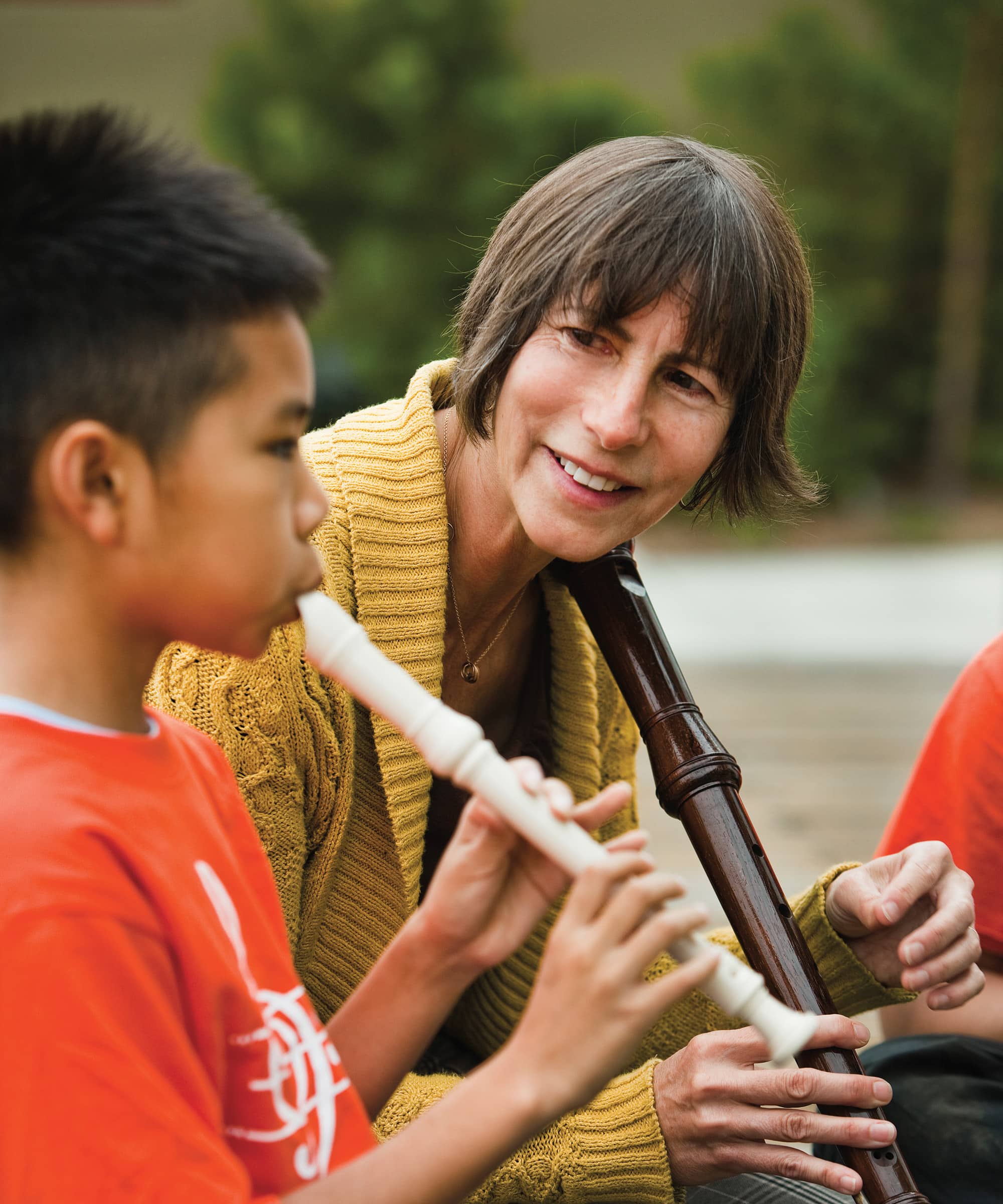 A person, with short dark hair wearing a yellow sweater holding an instrument, faces a child with short dark hair wearing a bright orange shirt playing the flute.