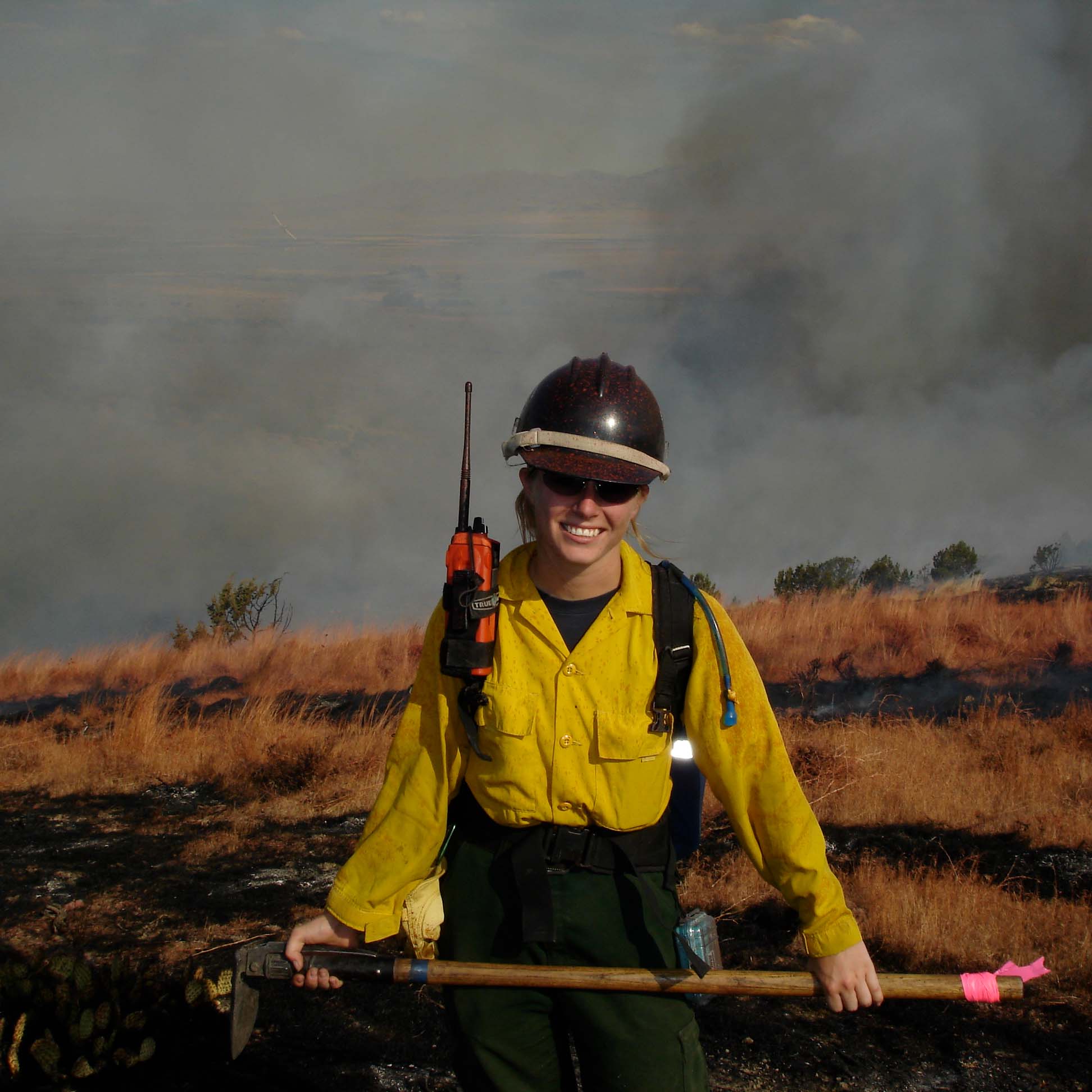 A person wearing a safety helmet wearing a yellow shirt and carrying a hoe smiles at the viewer in front of burnt vegetation.