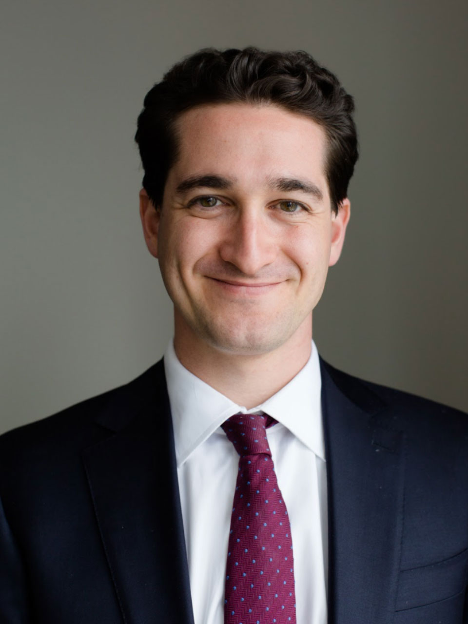 A person with short brown hair, a dark formal jacket, and a burgundy patterned tie smiles at the viewer.