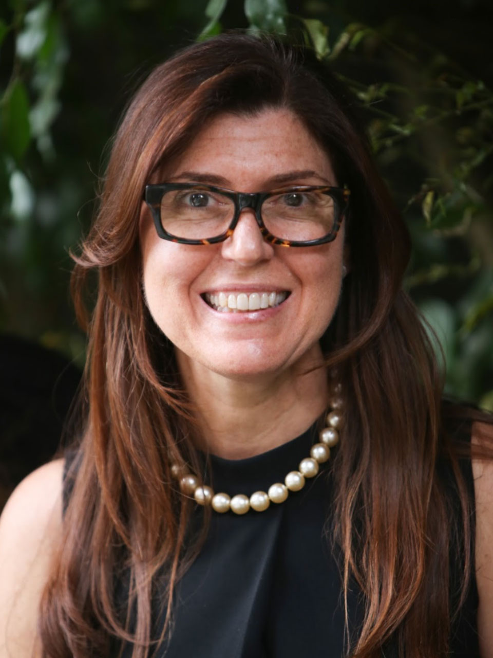 A person with long brown hair, glasses, and a dark sleeveless dress smiles at the viewer. The person is standing in front of a dark green bush.