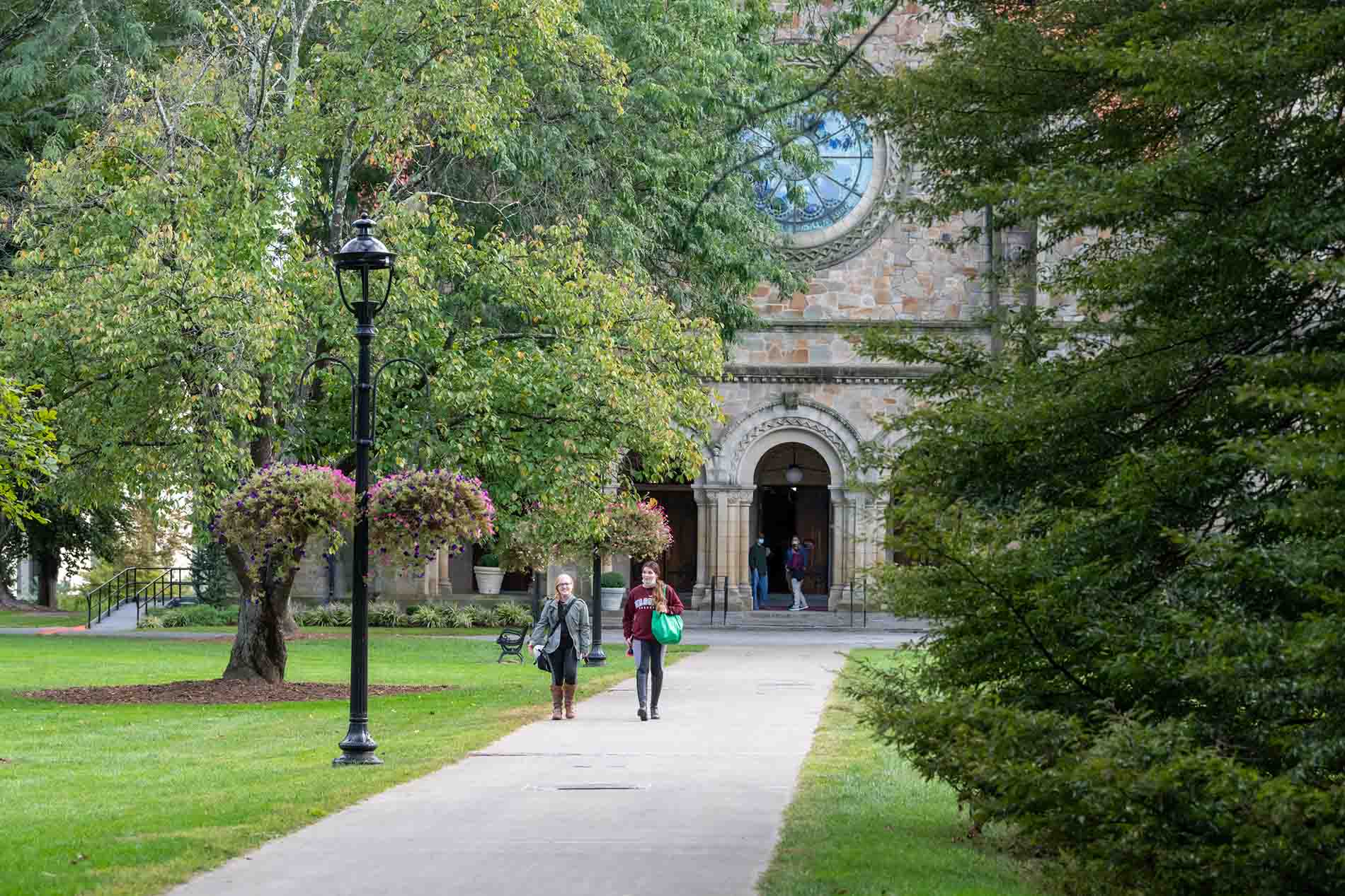 A photo of a straight walkway leading away from the chapel, a large, stone building with arches and a big round window. There are trees with green leaves on both sides of the walkway. It’s a sunny day. Two people, seen from a distance, are walking towards the viewer.
