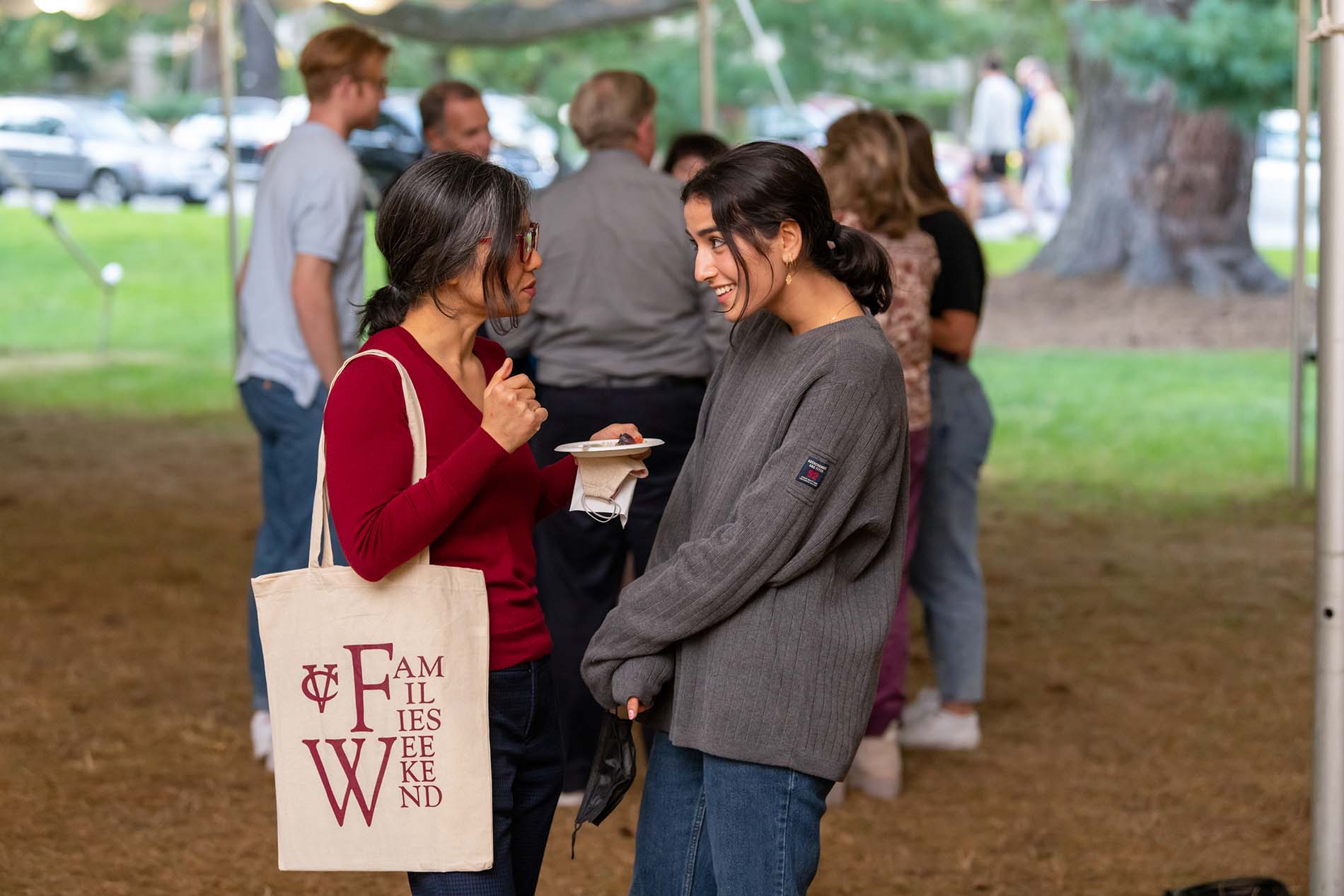 A parent and a child talk to each other while standing under a large event tent, face to face. The parent, on the left, has long, straight, black hair in a ponytail, glasses, a red shirt, and a bag with the text “Families Weekend” on it. The person on the right has long, black hair in a ponytail and a gray sweater. Blurred figures are visible in the background.