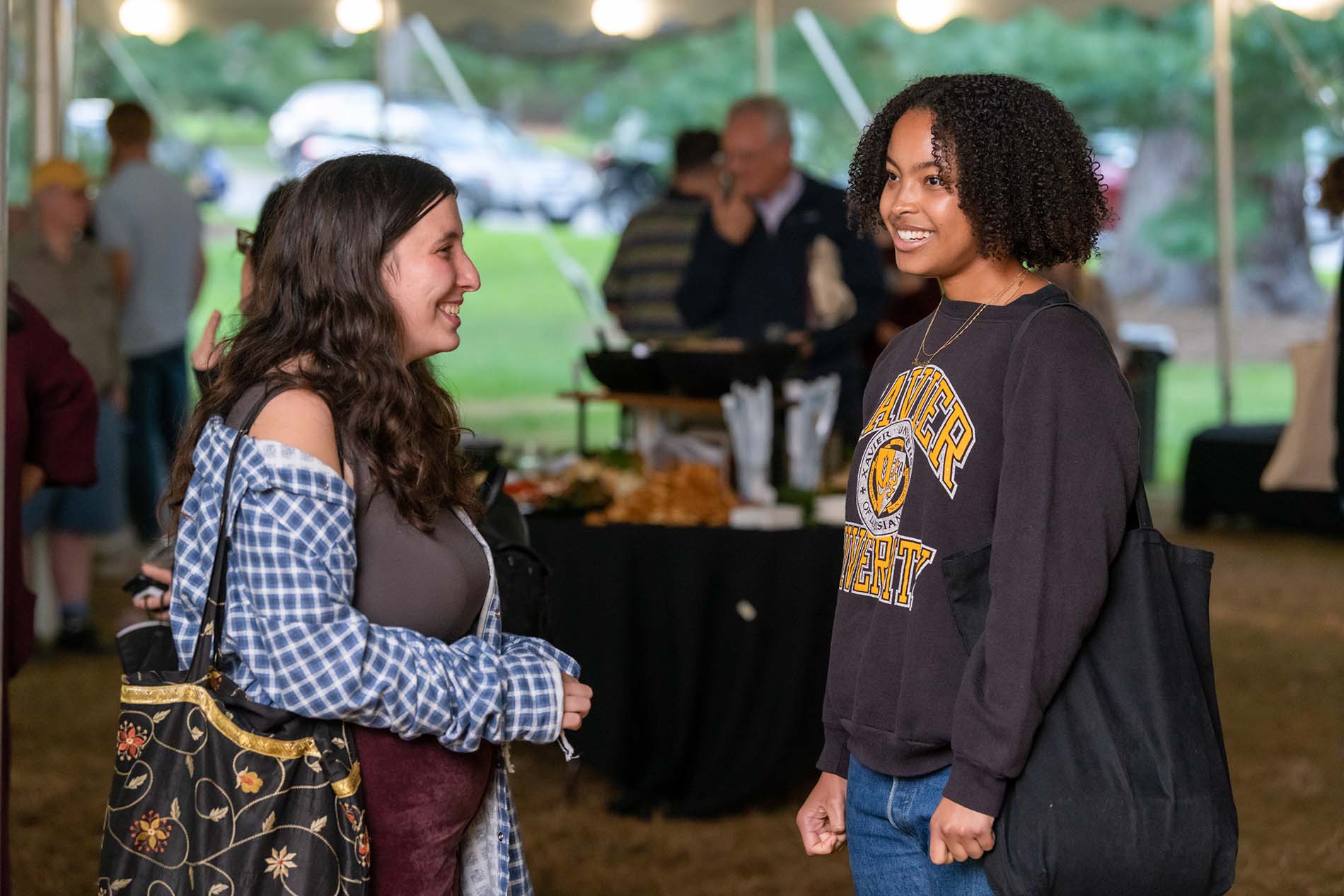 A photo of the opening reception for the weekend. Two people talk to each other while standing under a large event tent, face to face. The person on the left has long, straight, brown hair, a blue checkered shirt, and a flower-patterned bag. The person on the right has black, shoulder-length, curly hair, a brown sweater with an athletic logo on it, and a black shoulderbag. Blurred figures are visible in the background.