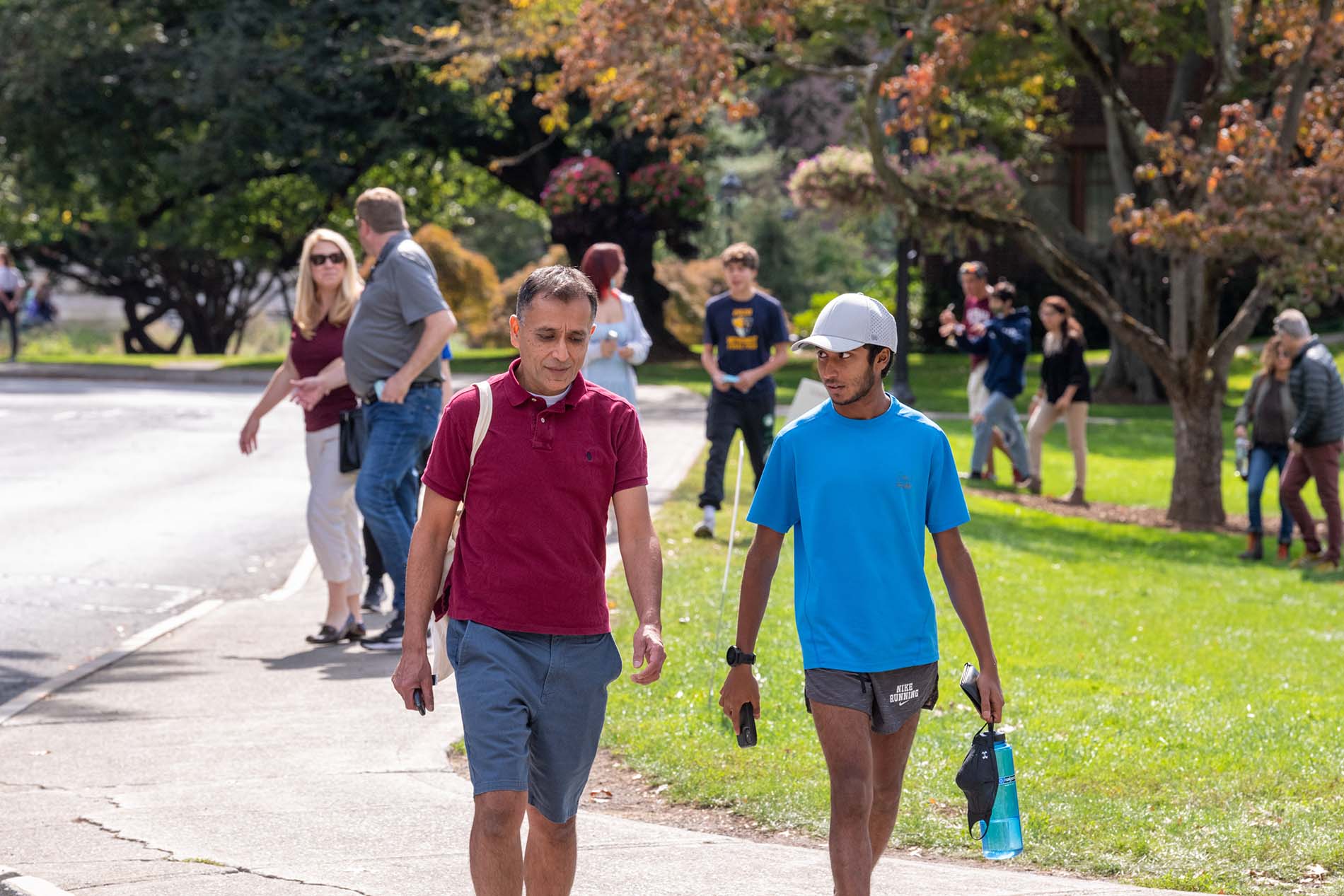 A parent and a child walk together on a sunny day. The parent, on the left, is wearing a maroon polo shirt and blue shorts. The child, on the right, is wearing a white baseball cap, a bright blue polo shirt, and gray shorts. Many people are visible walking in the background. There are a lot of trees.