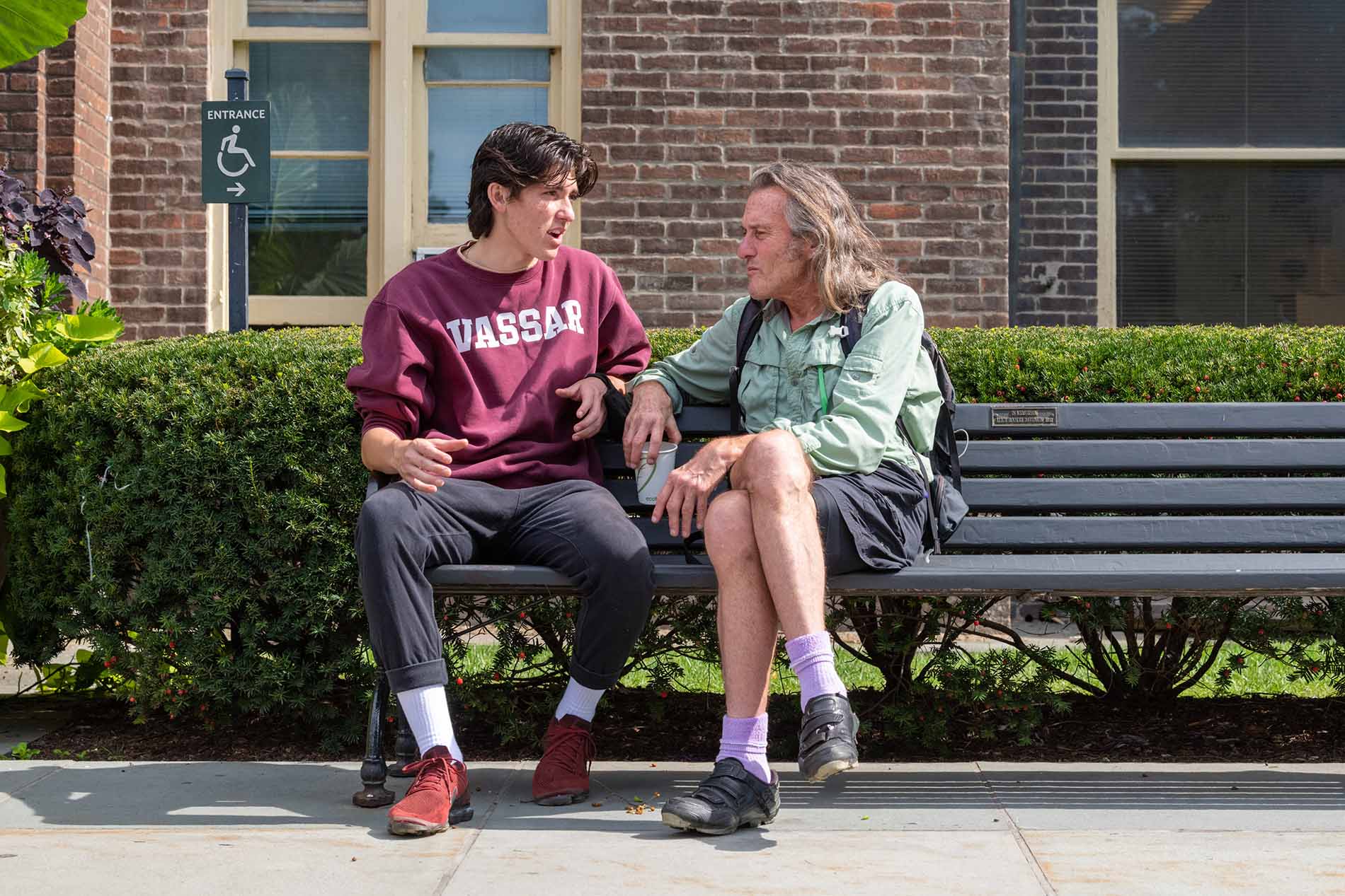 Two people, probably a child and a parent, sit on a bench outside. The child, on the left, has shortish, shoulder-length brown hair and a burgundy sweater with “Vassar” on it. The parent, on the right, has long graying hair, stubble, and a green shirt. There is a hedge behind the bench, and a brick building right behind the hedge.