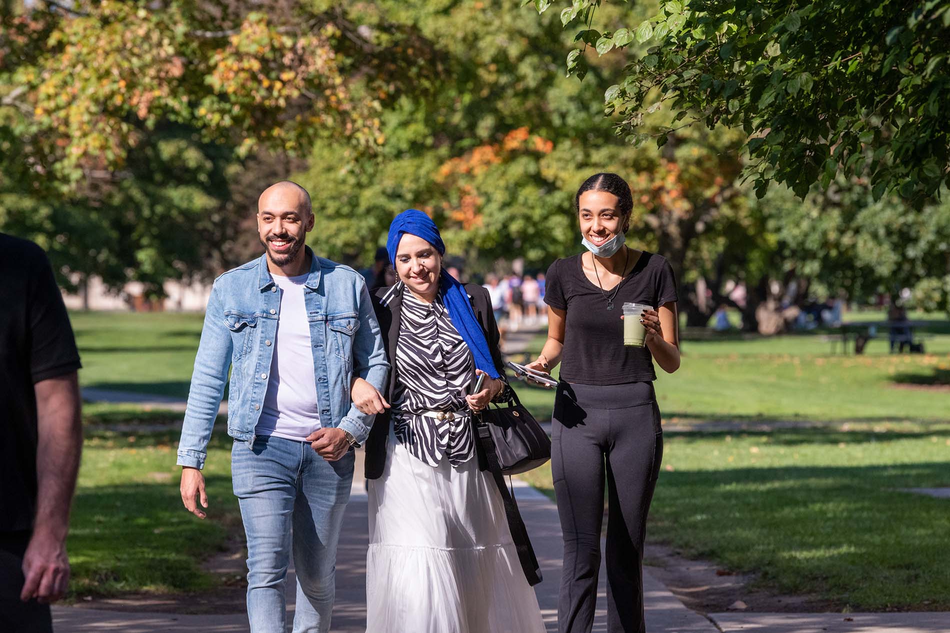 A family of three people walk together. The leftmost person has a shaved head, a beard, and a blue denim jacket. The middle person has a blue headscarf and a black-and-white striped shirt. The rightmost person has long, dark, curly hair and a black shirt. It is a sunny day. Trees with mostly green leaves are visible in the background.