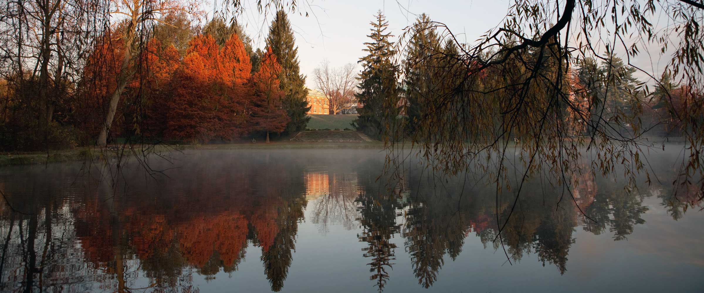 A photo of Sunset Lake in autumn. The lake is covered by fog, and the trees are brown with sunlight reflecting off them,