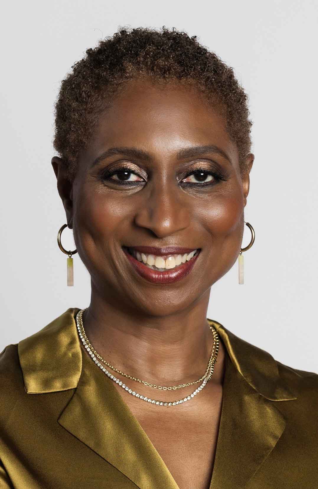 A portrait of a person with short, dark, curly hair and earrings smiling at the camera