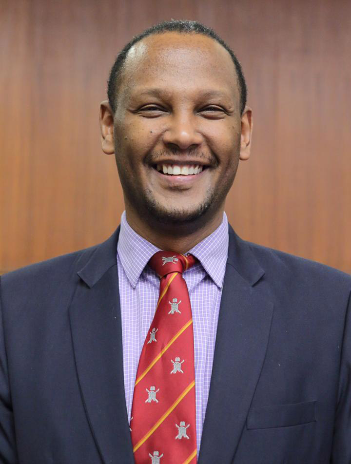 A headshot of Dr. Abebe Bekele, a person with short dark hair who is wearing a dark blue jacket, light purple patterned shirt and red tie.