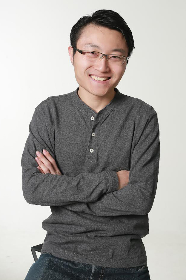 A person with shirt black hair, glasses, a gray button-down shirt, and dark jeans smiles at the viewer. The person has their arms folded across their chest.