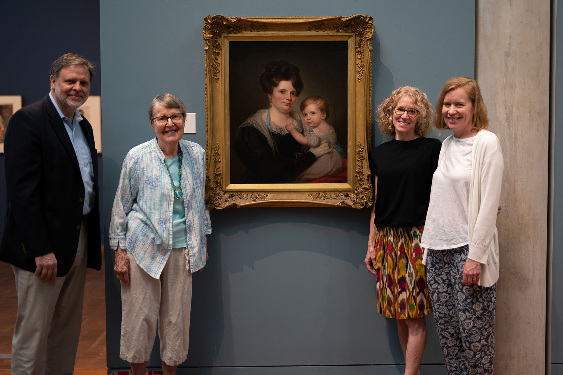 Four people standing next to a painting of a woman holding a child which is hanging on a blue-gray wall