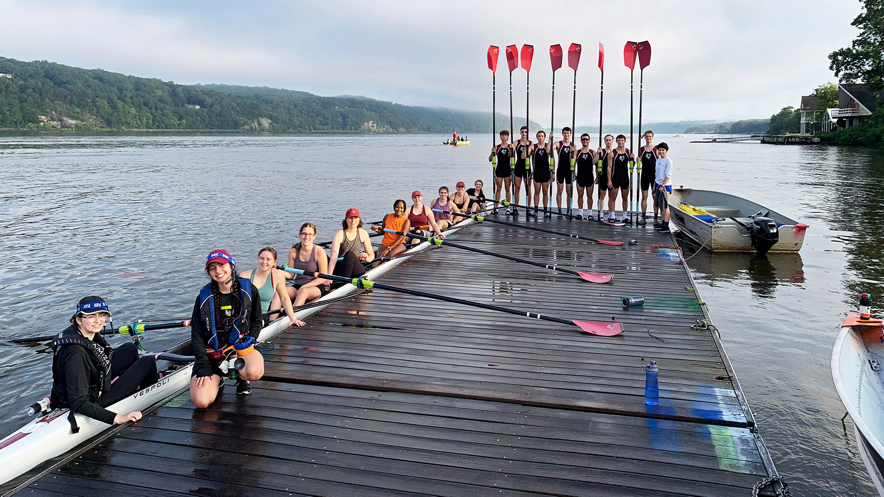 A dock on a lake with hills in the background. In the water a rowing scull is filled with people and five people on the dock holding a paddle.