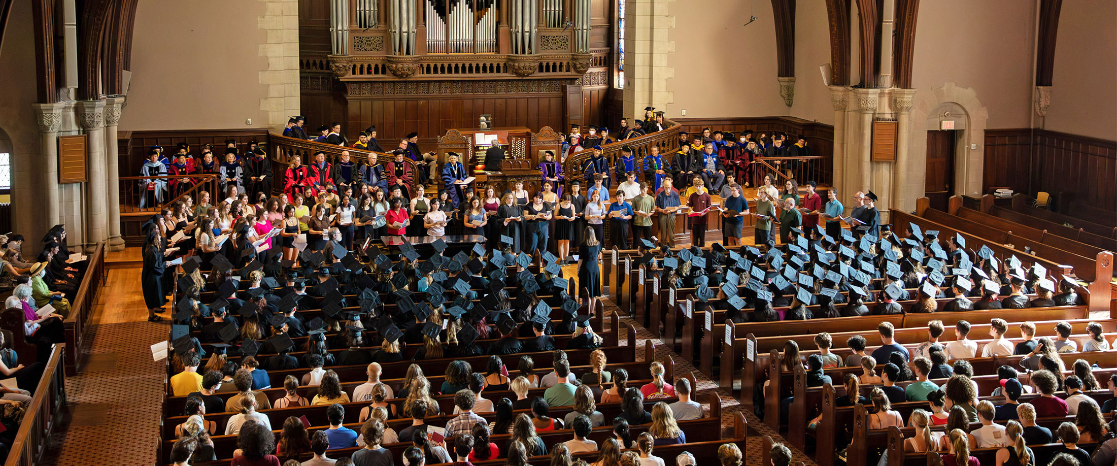 A large group of people sitting in a chapel. The people on stage are wearing different color graduation outfits while the people sitting at the pews are in traditional black graduation caps and gowns.