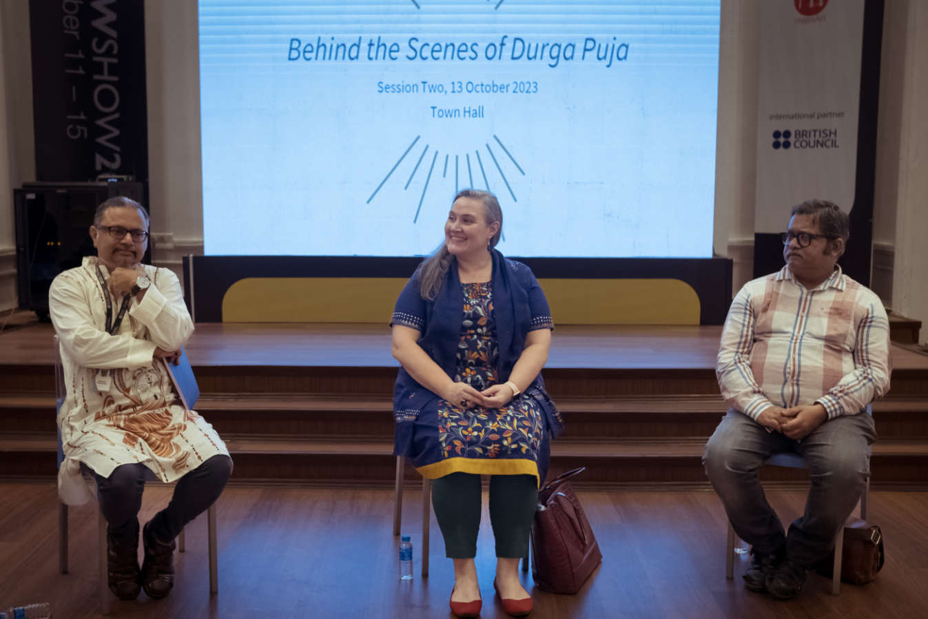 Three people sit in chairs onstage. Behind them is a screen showing a slide with the text “Behind the Scenes of Durga Puja, Session Two, 13 October 2023, Town Hall”.