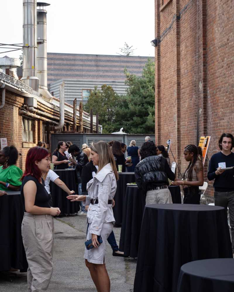 A gathering of people in an open space framed by brick industrial buildings. This is an event with small black cafe tables. People stand around the tables talking.