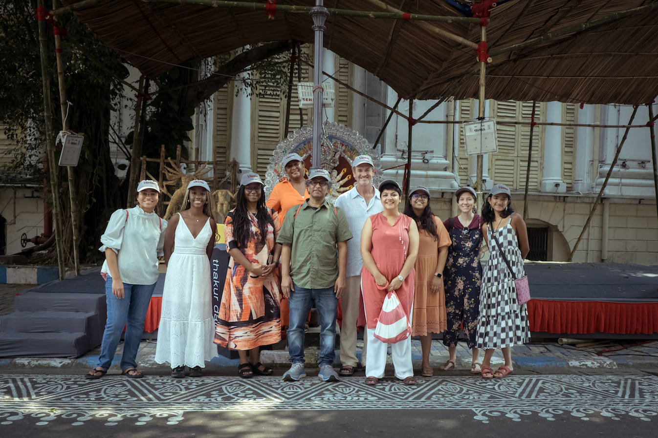 A group of people stand under a large straw roof, on a street with an intricate pattern painted on it. Behind them is a large stone building.