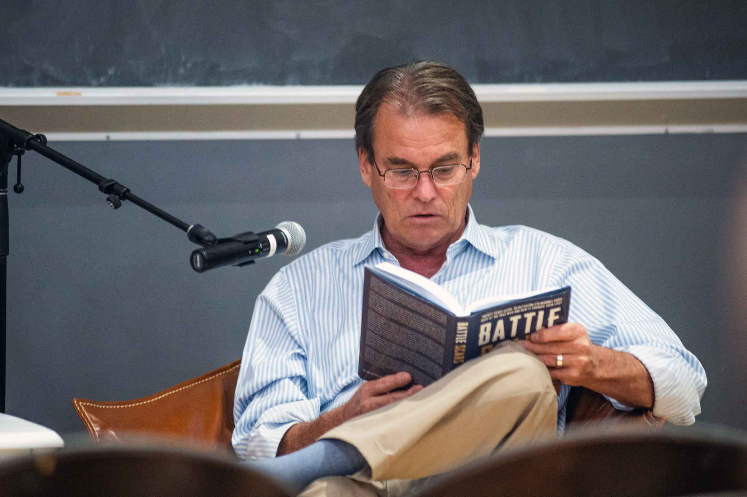 A person with glasses seated behind a microphone reading from a book with the work, Battle, and the cover.