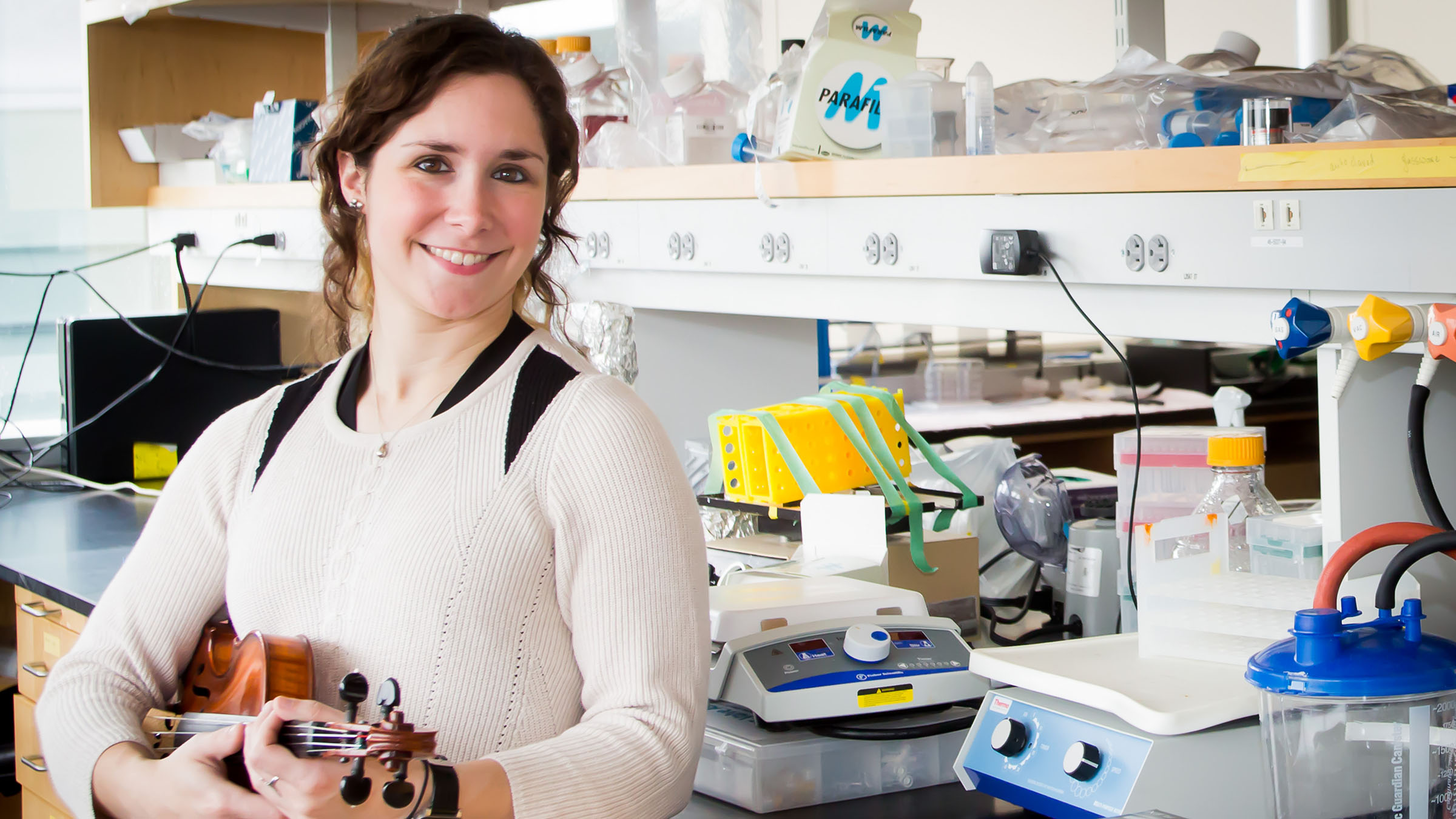 A person with long brown hair smiling at the camera while holding a violin in a laboratory filled with scientific equipment. 