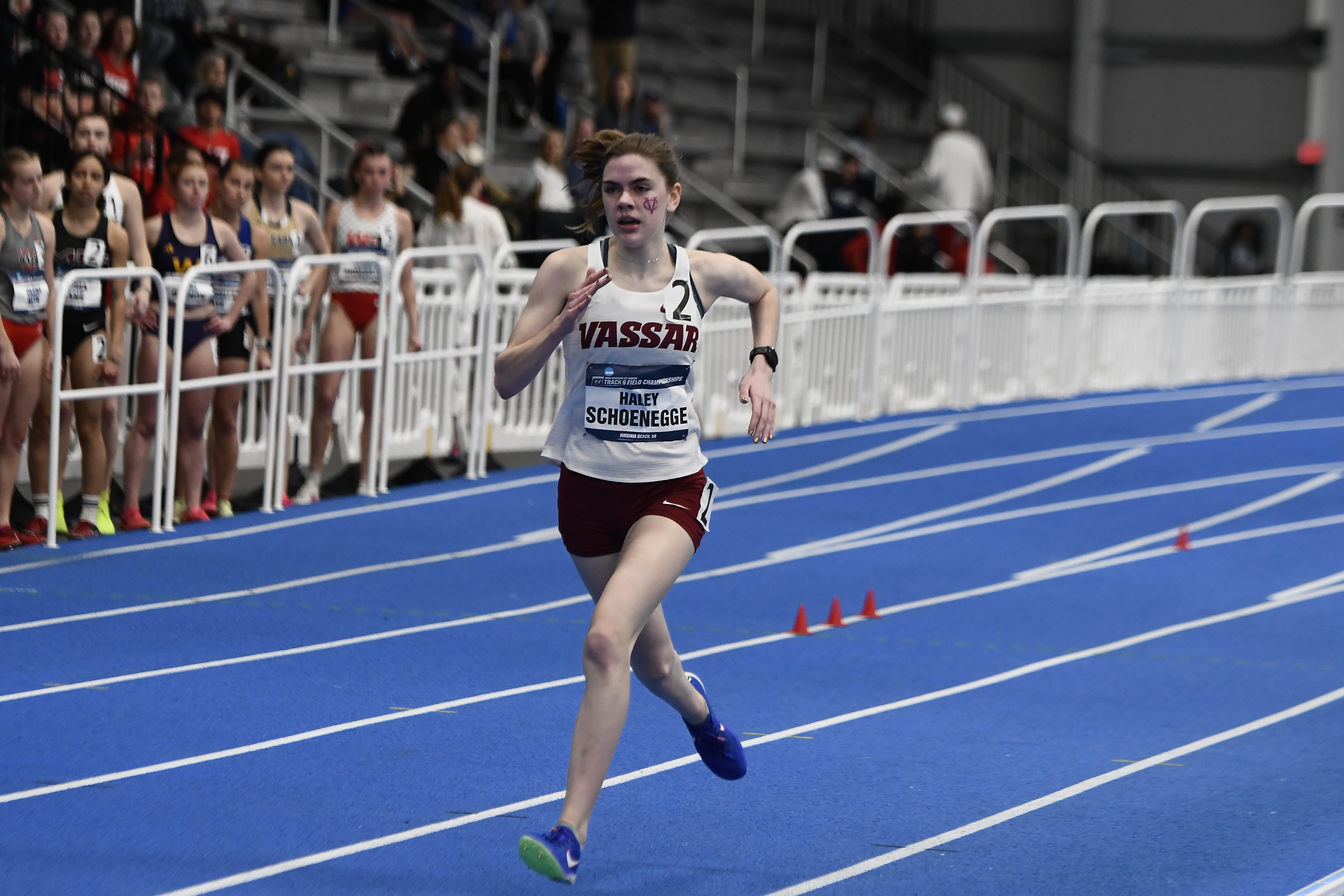 A person with a white Vassar tank top and burgundy shorts runs on a track. The person is wearing a tag with "2" on it, and another with "Haley Schoenegge" on it.